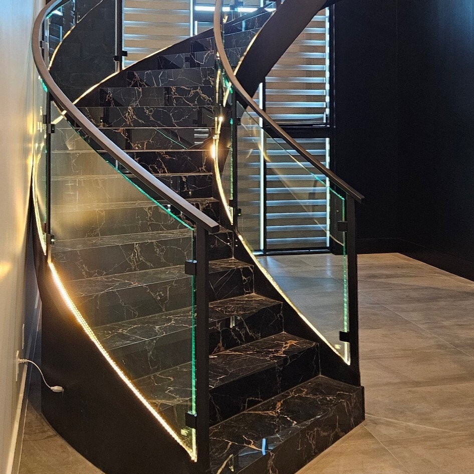 Curved glass railing with a black handrail following a spiral staircase with lit marble steps