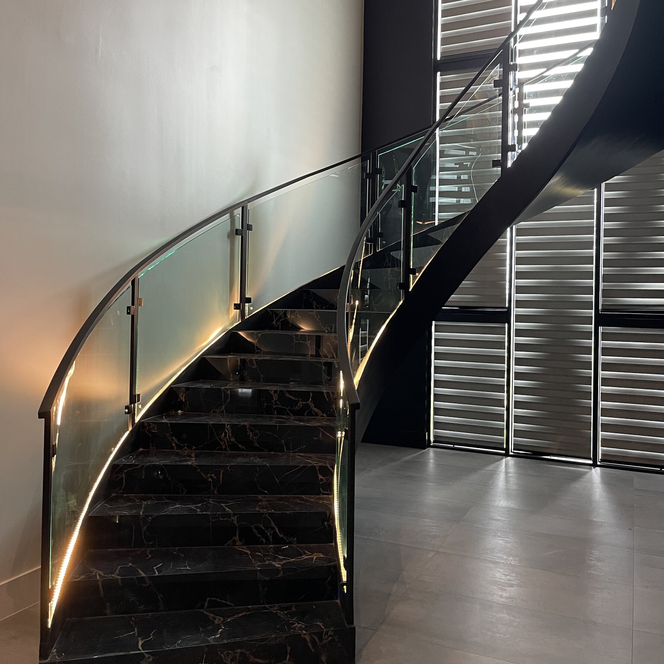 Curved frameless glass stair railing rising along a black marble spiral staircase