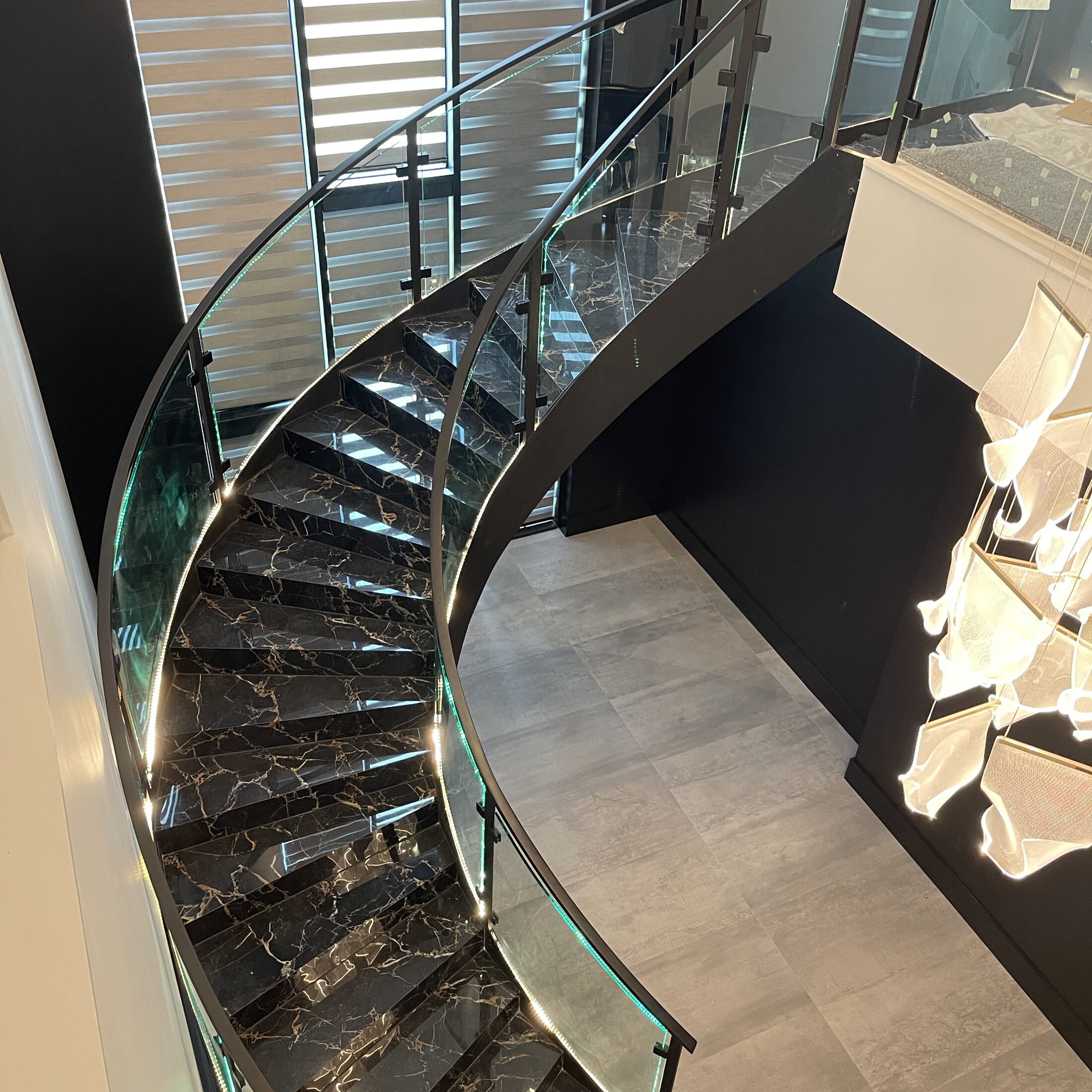 View down a spiral staircase lined with curved glass railing panels and a black handrail