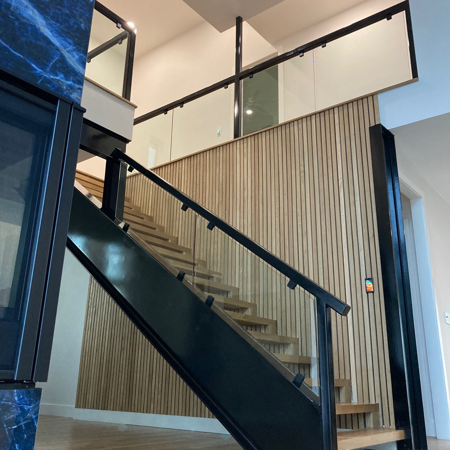 Glass stair railing with a black handrail on an open staircase beside a wood slat wall