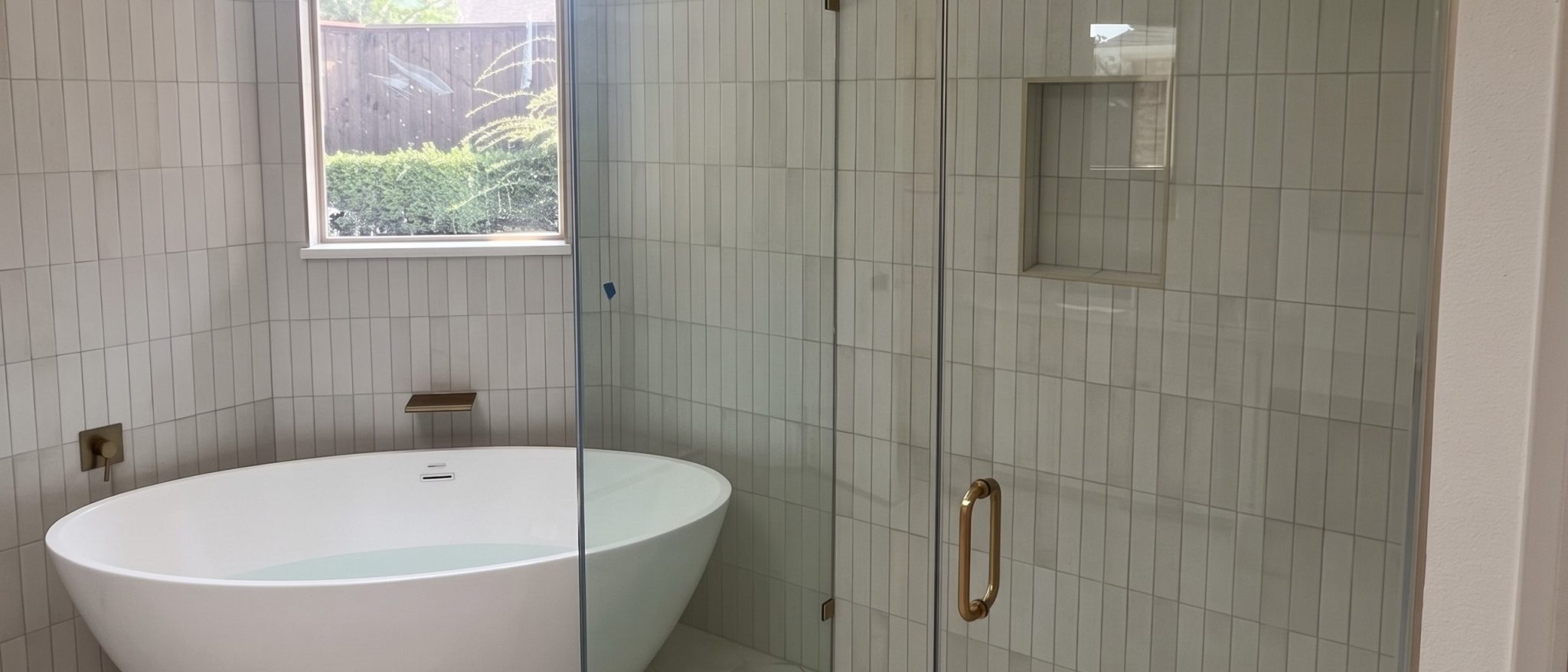 Frameless 90 degree glass shower enclosure with a satin brass pull beside a freestanding tub