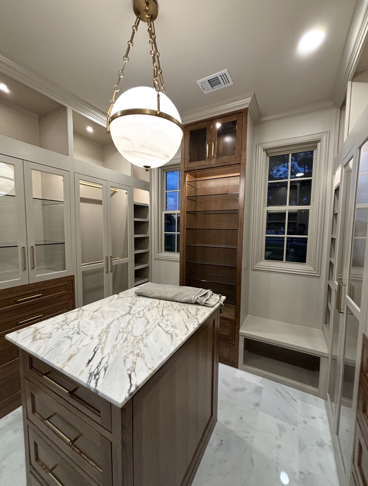 Walk-in closet with custom clear glass cabinet inserts and a marble-top island in Bartonville, TX